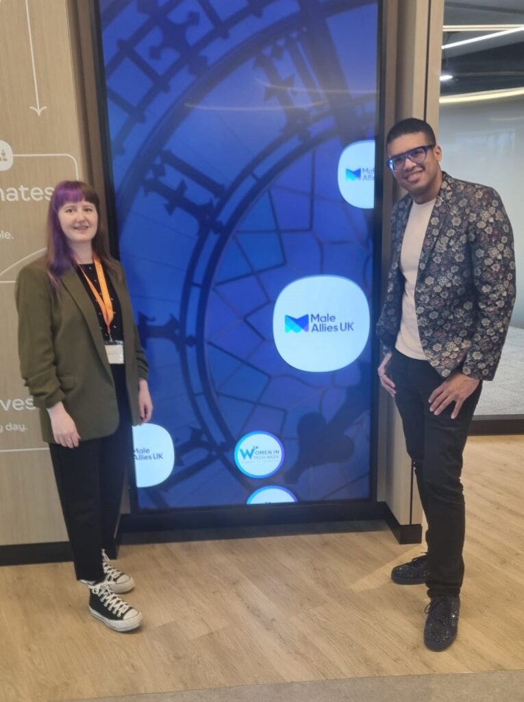 Jade and Lee are stood either side of a tall, thin screen that has bubbles going across it with the Male Allies UK logo in them. Jade's wearing a khaki blazer and Lee's wearing a blue and silver floral blazer and they're smiling at the camera.