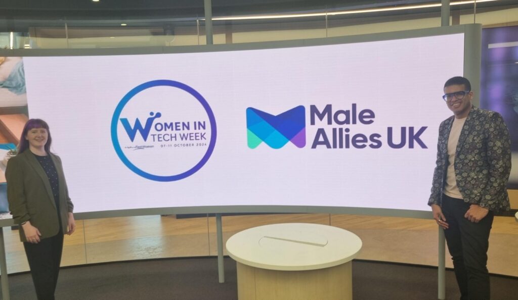 Jade is wearing a khaki blazer and Lee is wearing a blue and silver floral blazer. They're standing either side of a large curved screen that has the Male Allies UK and Women In Tech Week logos on it.